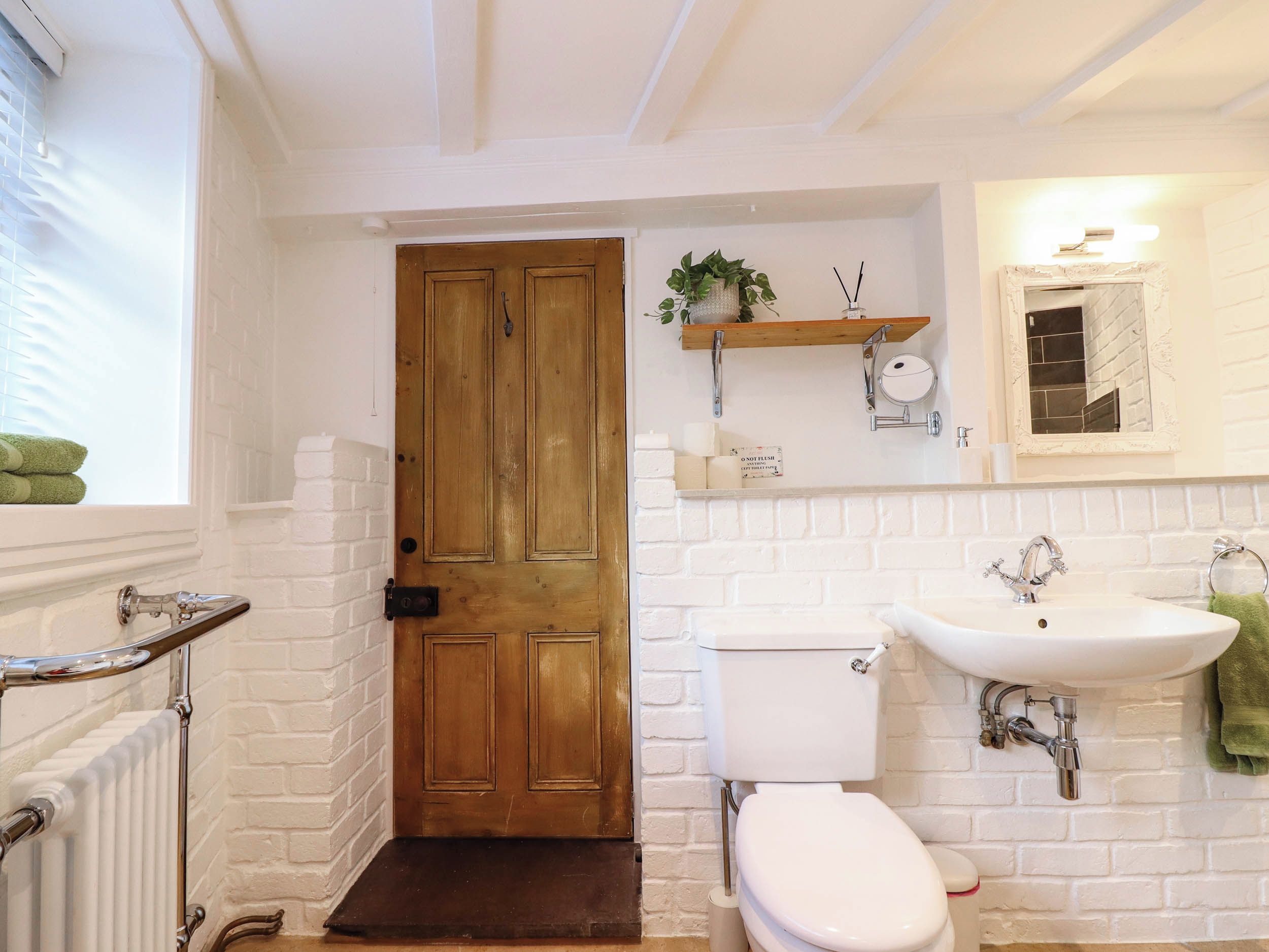 Une salle de bain avec un mur en briques blanches, une porte en bois, des toilettes blanches, un lavabo avec un miroir et une étagère contenant des articles de toilette.