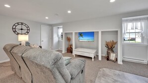 Una sala de estar con un sofá gris, una mesita de centro blanca, un televisor montado en la pared y un reloj.