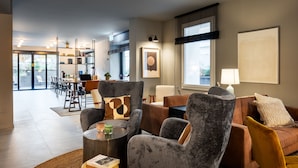 Una sala de estar moderna con mobiliario tapizado en tonos grises y marrones, una zona de comedor con una mesa y sillas, y un ventanal con persianas.