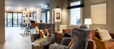 A modern living room with grey and brown upholstered furniture, a dining area with a table and chairs, and a large window with blinds.