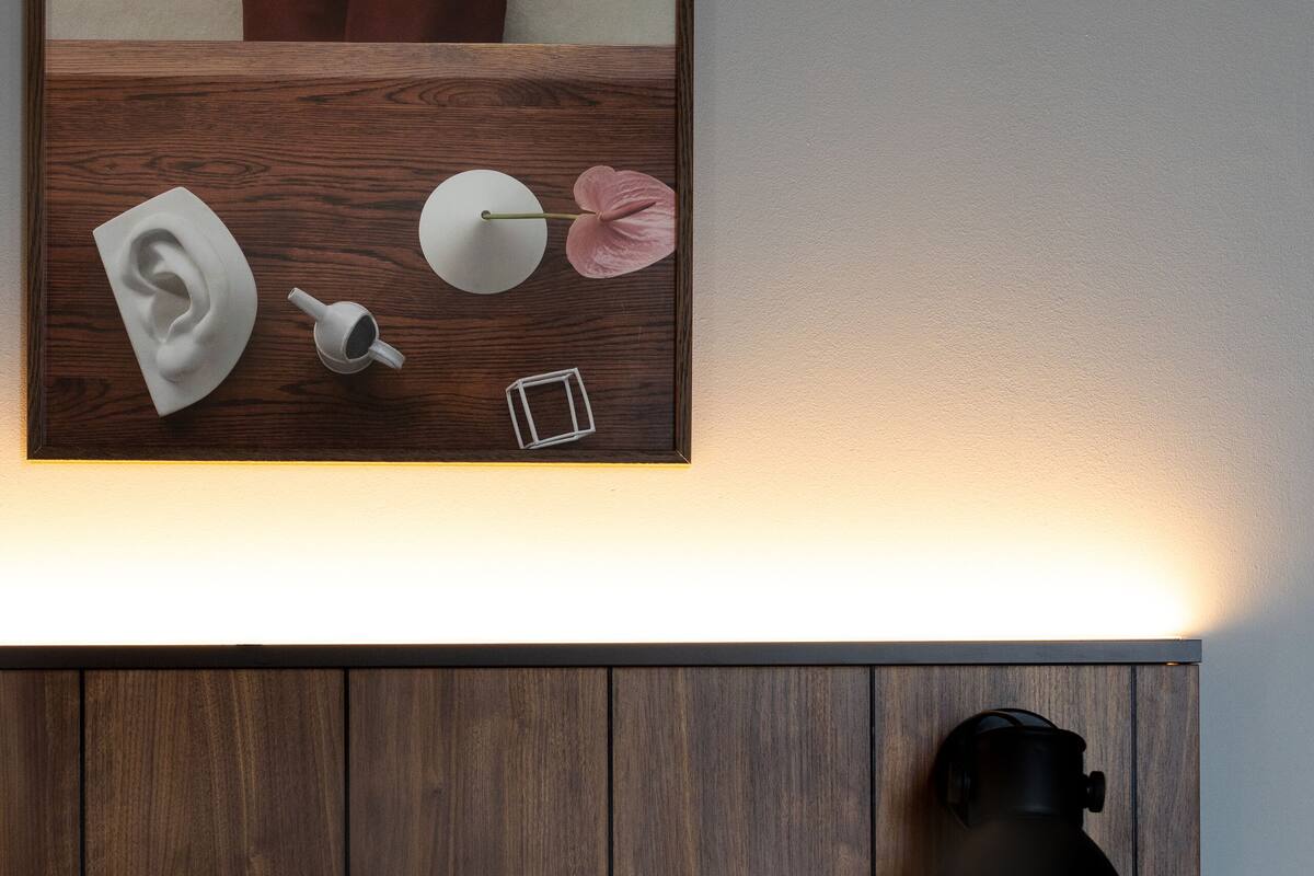 A neatly made bed with white linens, a bedside table with a lamp, a small plant, and a wall-mounted control panel.