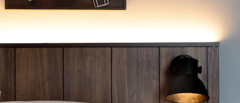 Un lit bien fait, avec du linge de lit blanc, une table de chevet avec une lampe, une petite plante et un panneau de commande fixé au mur.