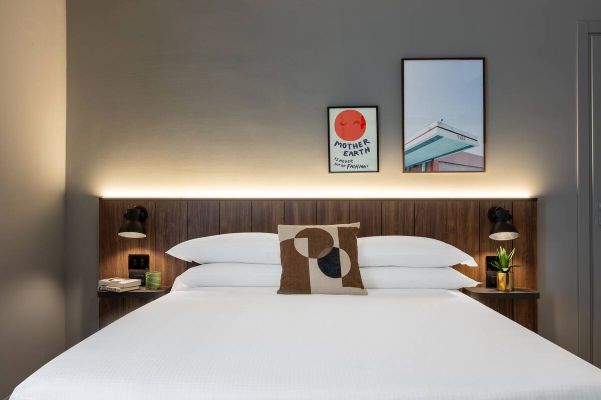A neatly made bed with a pillow, two bedside tables with lamps, a framed picture on the wall, and a small plant on one of the tables.