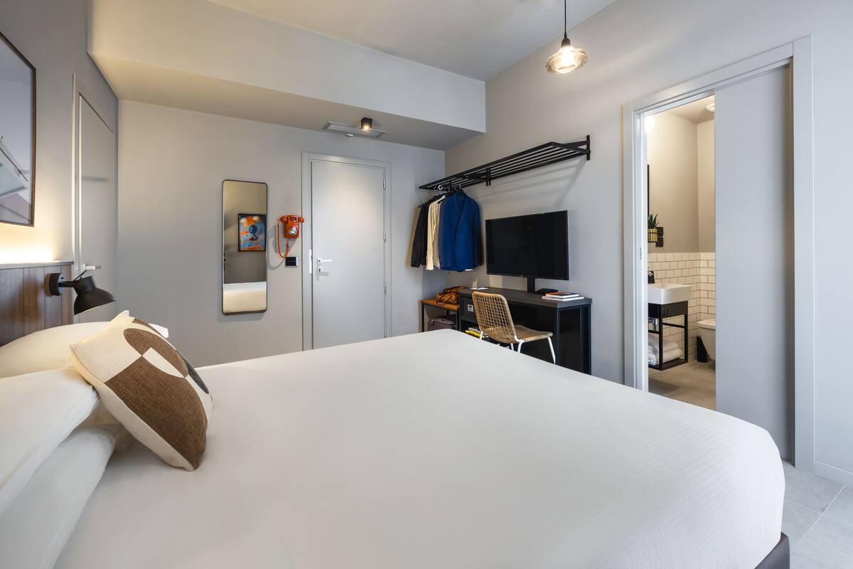 A modern hotel room with a large bed, a desk with a computer, a chair, a TV, and a bathroom visible through an open door.
