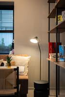 Una sala de estar moderna con un sofá, un otoman negro redondo, una lámpara de pie y una estantería con libros y una caja.