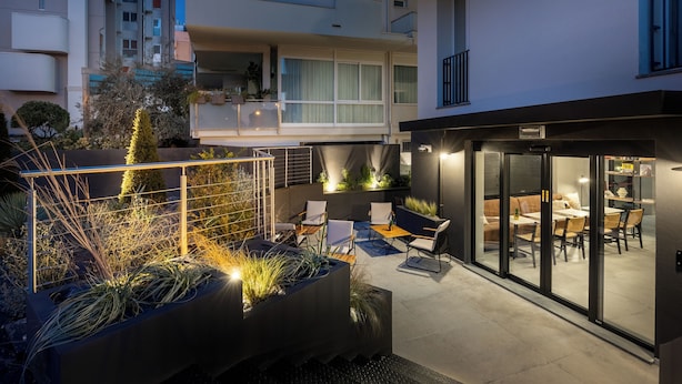 Un patio exterior moderno con mobiliario, una entrada con puerta de vidrio y un interior bien iluminado visible a través del vidrio.