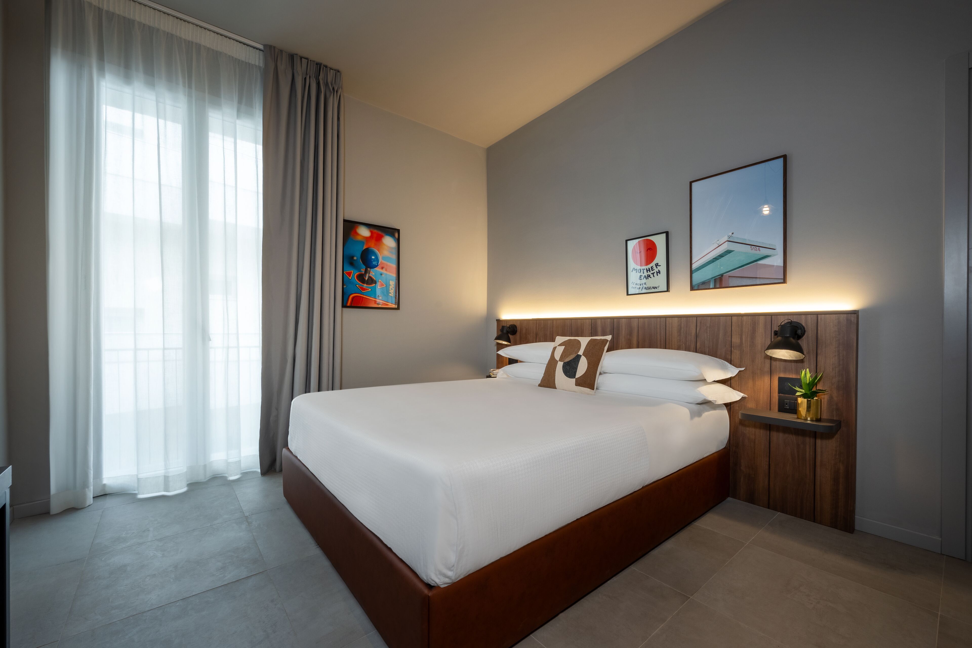 Moderni hotellihuone, jossa on suuri sänky, yöpöytä lampun kanssa ja taideteoksia seinillä.