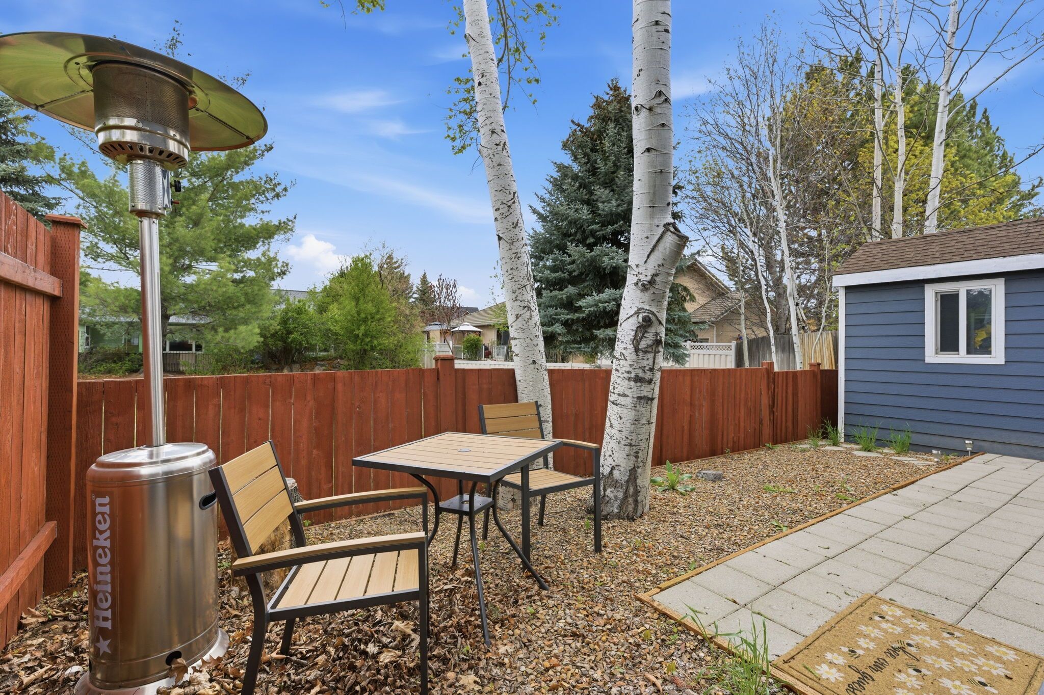 A backyard with a patio table and chairs, a Heineken branded heater, a wooden fence, and a small shed.
