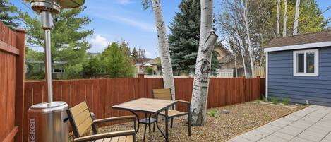 A backyard with a patio table and chairs, a Heineken branded heater, a wooden fence, and a small shed.