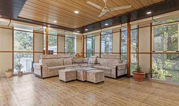 A spacious living area with a sectional sofa, a glass coffee table, and a ceiling fan. Large windows provide views of the outdoors. - Treebo Olive Lake View Inn June Estate Bhimtal (Nainital)