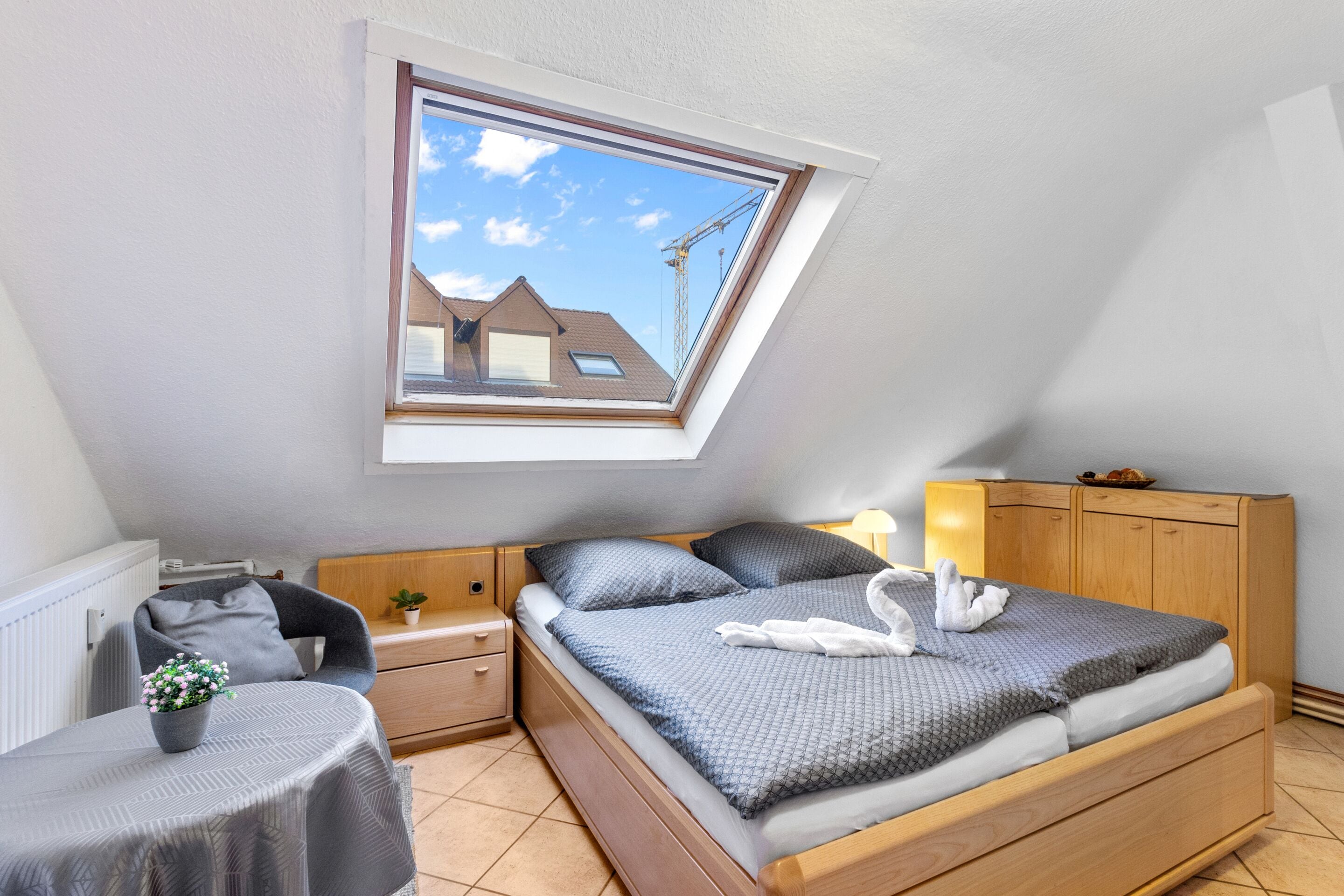 A bedroom with a skylight, a bed with a grey bedspread, a bedside table with a plant, and a wooden wardrobe.