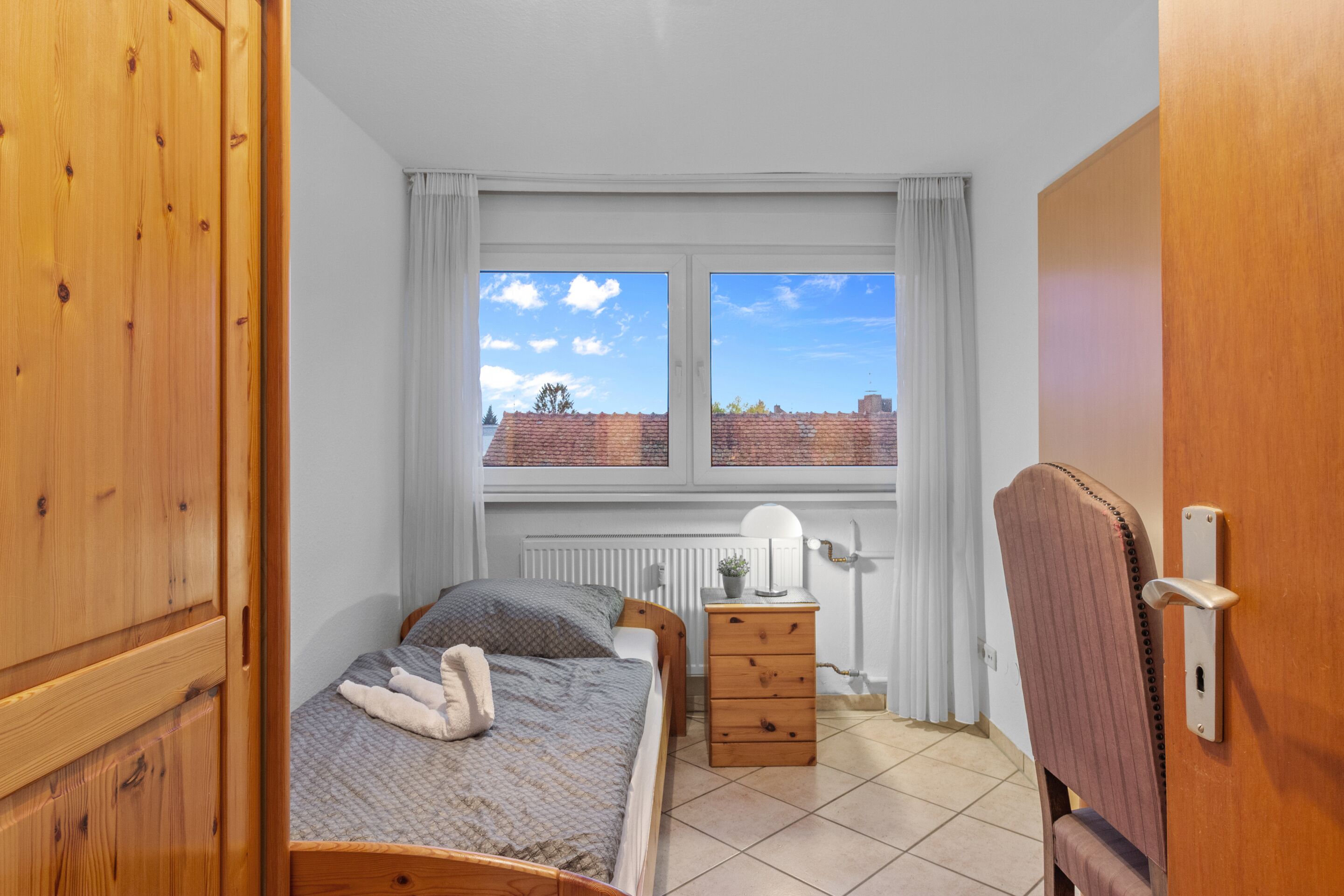 Een slaapkamer met een bed, een houten dressoir, een stoel en een raam met uitzicht op gebouwen.