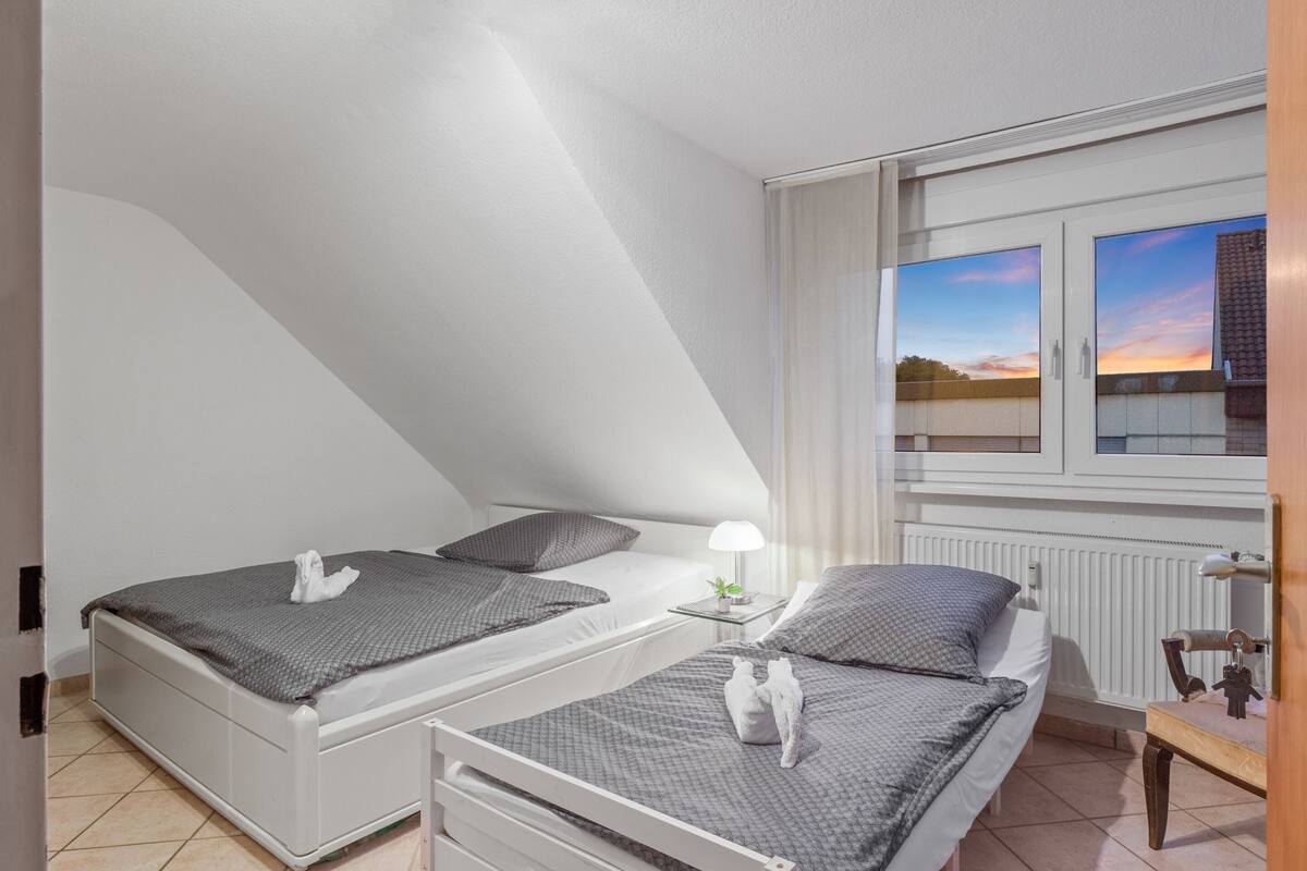 A bedroom with two beds, a nightstand, a lamp, and a window with a view of the sky.