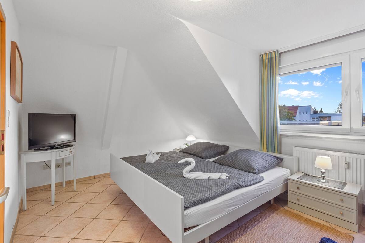 A bedroom with a bed, a TV on a stand, a bedside table with a lamp, and a window with curtains.