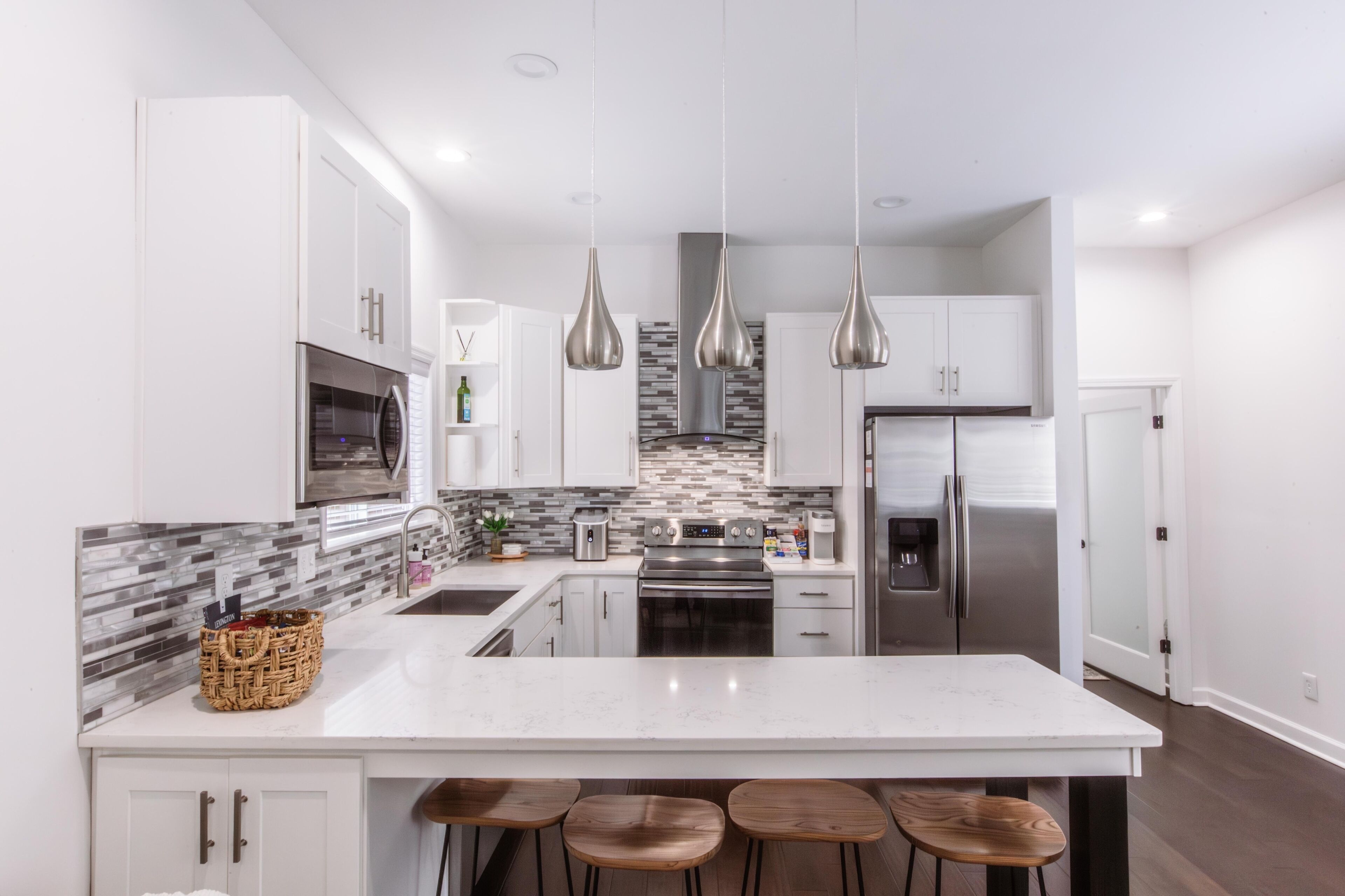 Une cuisine moderne avec des meubles blancs, des appareils en acier inoxydable, un îlot en marbre et des luminaires suspendus.
