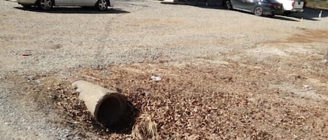 Un estacionamiento con grava y un tubo grande al frente, un edificio con una escalera en el fondo, y coches estacionados cerca.