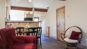 A cozy living room with a red sofa, a round wicker chair, and a dining table with chairs. A wooden door and a staircase are visible in the background. - Bluebird Day - Casa Sol: Cozy 2BR townhome in Bend, OR, featuring two King beds and a hot tub. (Bend)