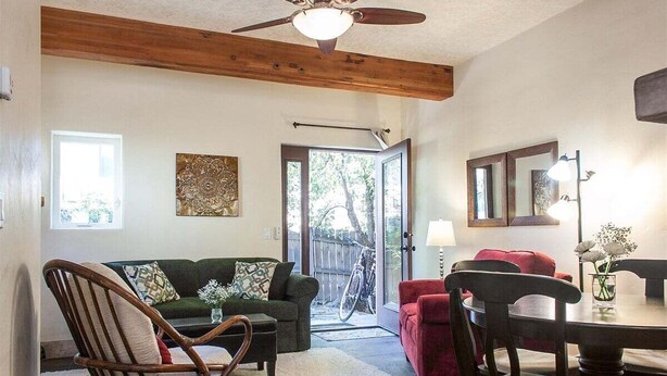 A living room with a wooden ceiling, a sofa, a chair, a dining table with chairs, and a door leading outside. - Bluebird Day - Casa Sol: Cozy 2BR townhome in Bend, OR, featuring two King beds and a hot tub. (Bend)