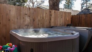 Un jacuzzi en un patio trasero con valla de madera, un árbol y una casa al fondo.