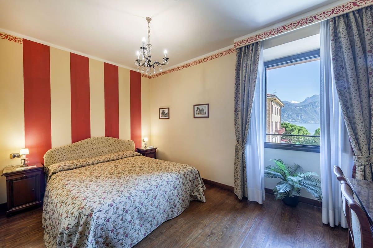 A hotel room with a bed, a nightstand, a chandelier, a window with curtains, and a view of a mountain.