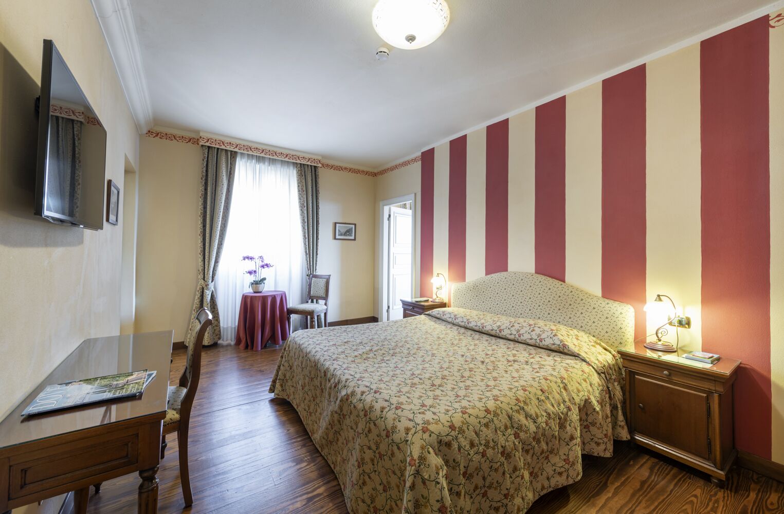 Ein Schlafzimmer mit einem Bett, einem Schreibtisch, einem Stuhl, einem Tisch mit Blumen und einem Fernseher an der Wand.