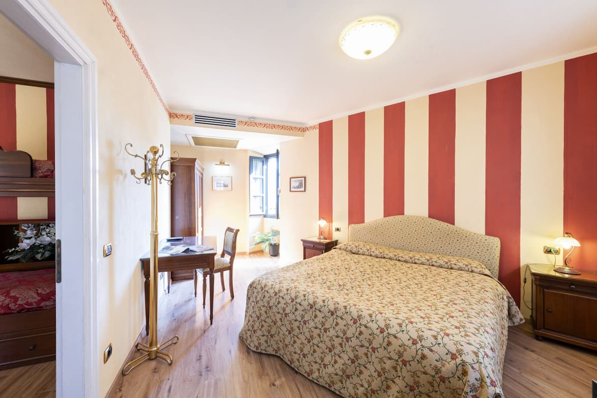 A bedroom with a bed, a desk, and a chair. There is a window with curtains, a radiator, and a lamp on the bedside table.