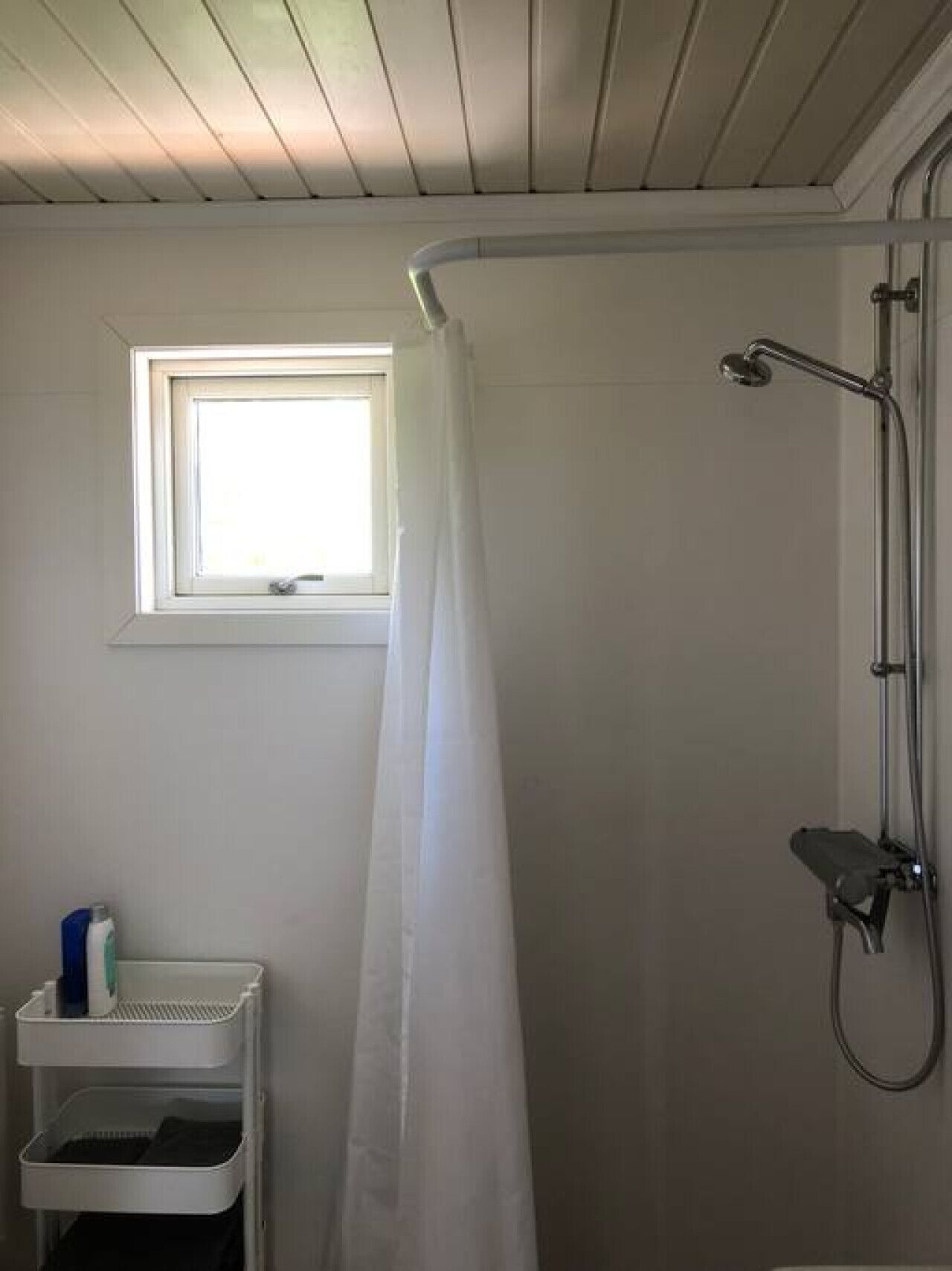 Et badeværelse med bruser, et vindue og en hvid hylde med toiletartikler.