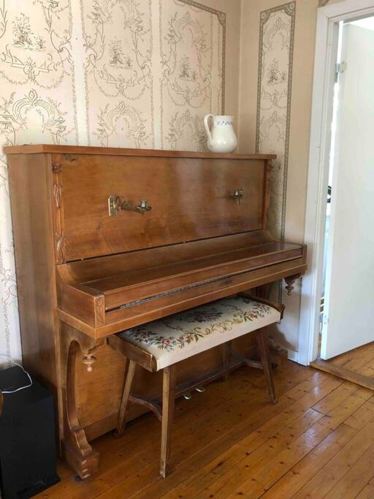 A wooden piano with a bench, a white pitcher on top, and patterned wallpaper.