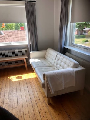 A room with a white sofa, a radiator, a window with curtains, and a view of a red-tiled roof outside.