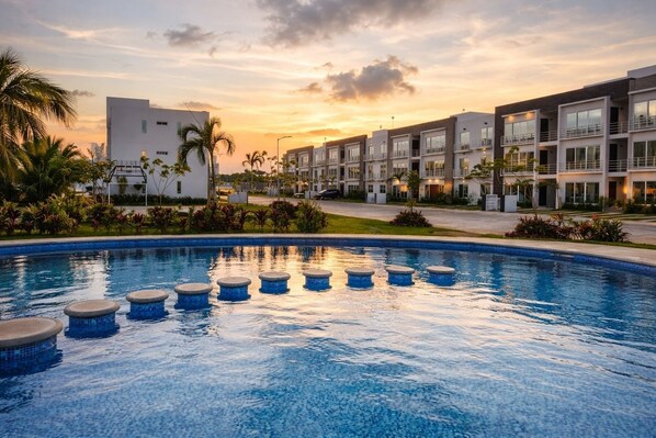 A swimming pool with circular steps, surrounded by modern buildings and palm trees, during sunset. - Nvo. Dept 3 Bedrooms with pool 18 min from beaches (Cancún)