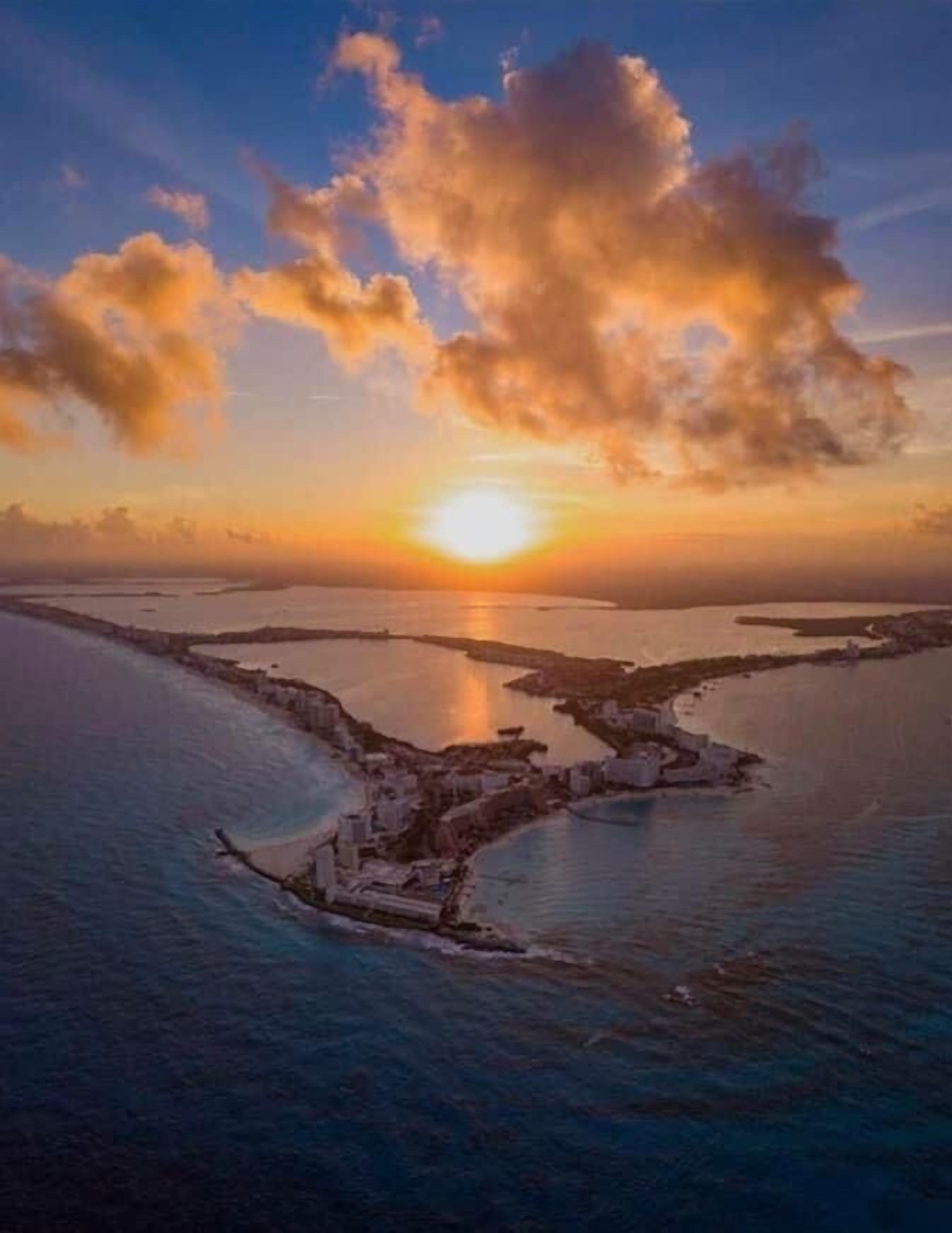 An aerial view of a coastal area with a sunset, featuring a large body of water, a small island with buildings, and a clear sky.