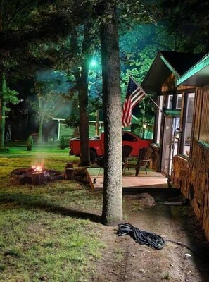 A rustic cabin with a fire pit, an American flag, and a red vehicle parked nearby. - Spohn-taneous Retreat (Grove)