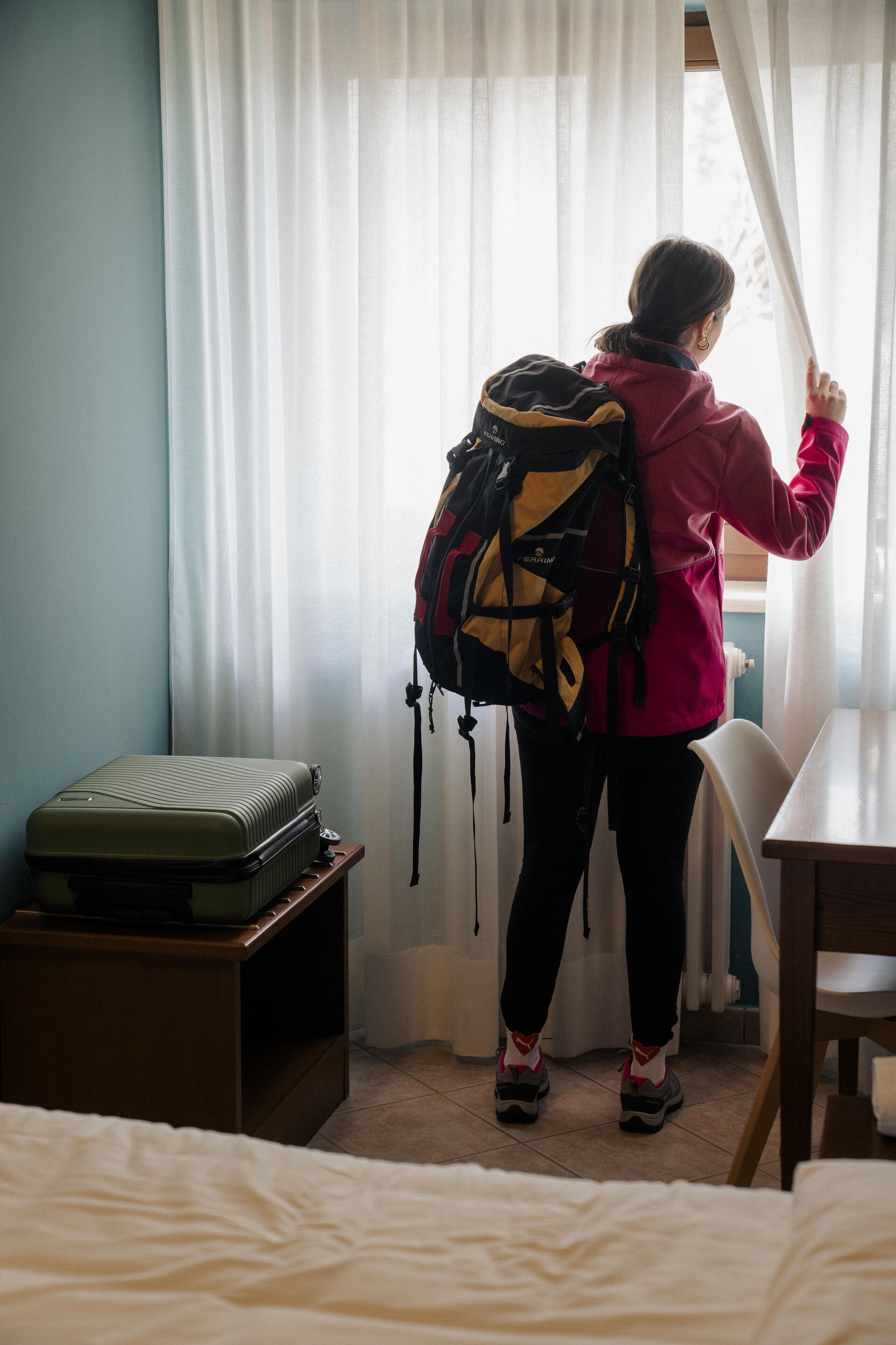 Una persona con una mochila de pie junto a una ventana con cortinas, una maleta sobre una mesa y una cama en primer plano.