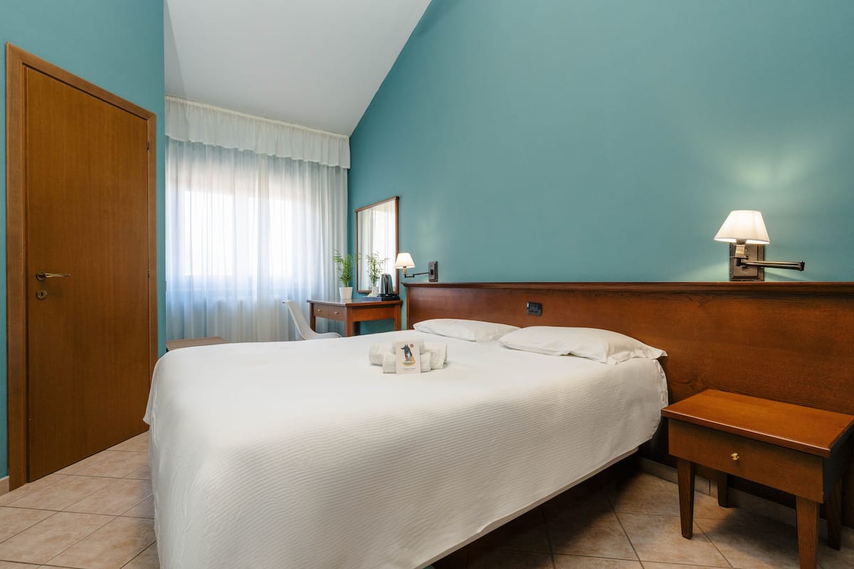 A hotel room with a large bed, a bedside table with a lamp, a wooden nightstand, and a mirror on the wall.
