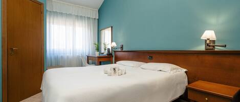 A hotel room with a large bed, a bedside table with a lamp, a wooden nightstand, and a mirror on the wall.