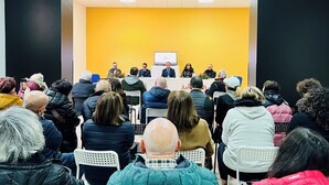 Un salón de conferencias con un panel de oradores en una mesa, público sentado frente a ellos, y una pared amarilla al fondo.