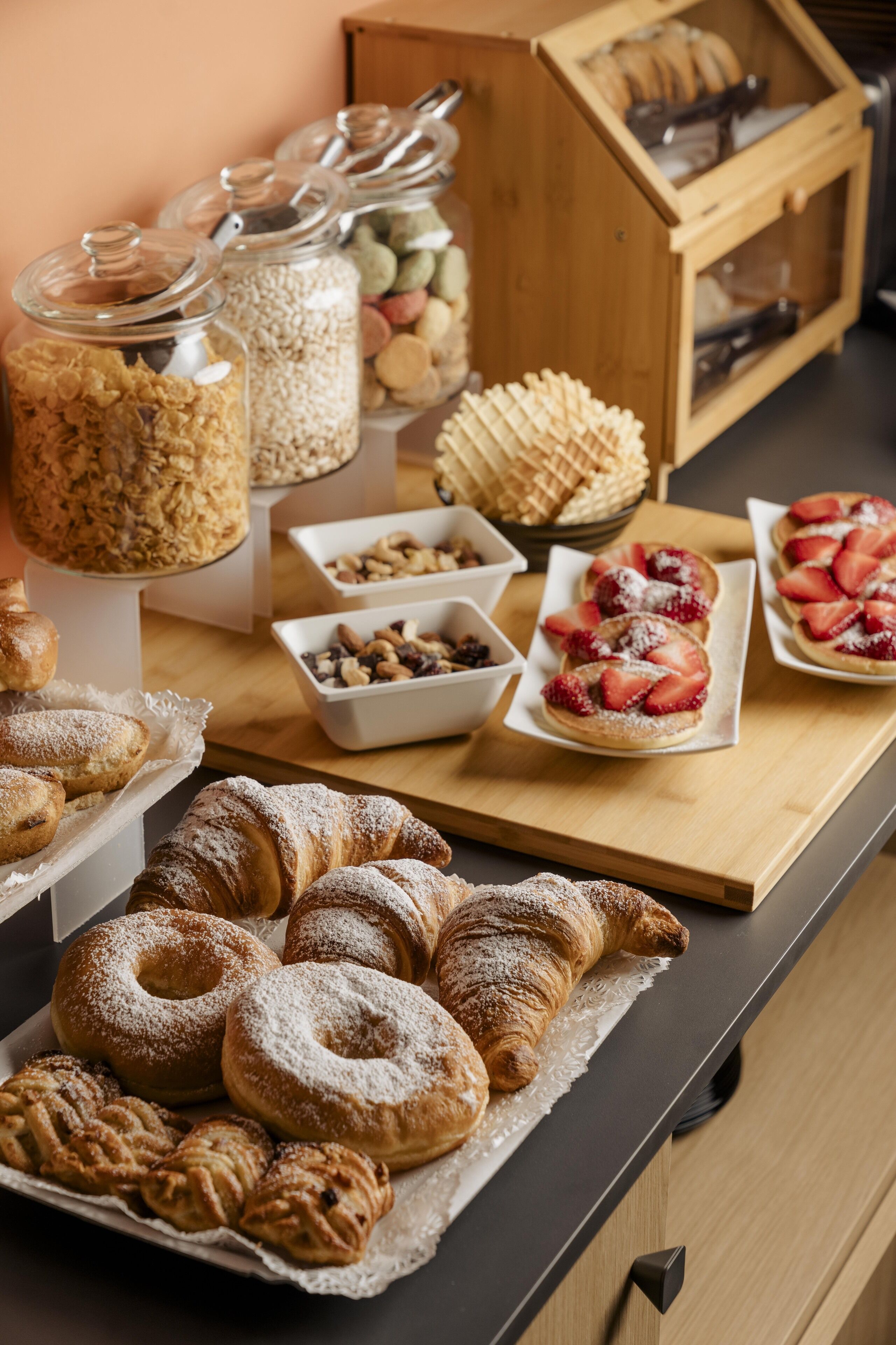 Una mesa de buffet con diversas pastas, incluyendo cruasanes y postres con fruta, y un mueble de madera con botes de nueces y semillas.