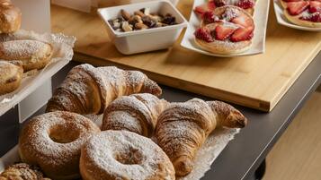 A buffet table with various pastries, including croissants and fruit-topped desserts, and a wooden cabinet with jars of nuts and seeds.
