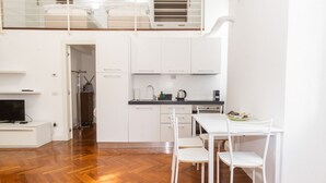 Una cuina compacta amb armaris blancs, una taula de dinar i cadires. Una televisió reposa sobre un moble a l'esquerra.
