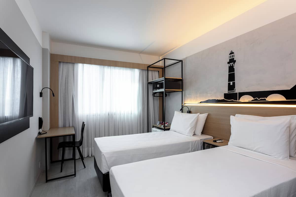 A hotel room with two beds, a desk, a chair, a TV, and a wall-mounted shelf with a lamp.