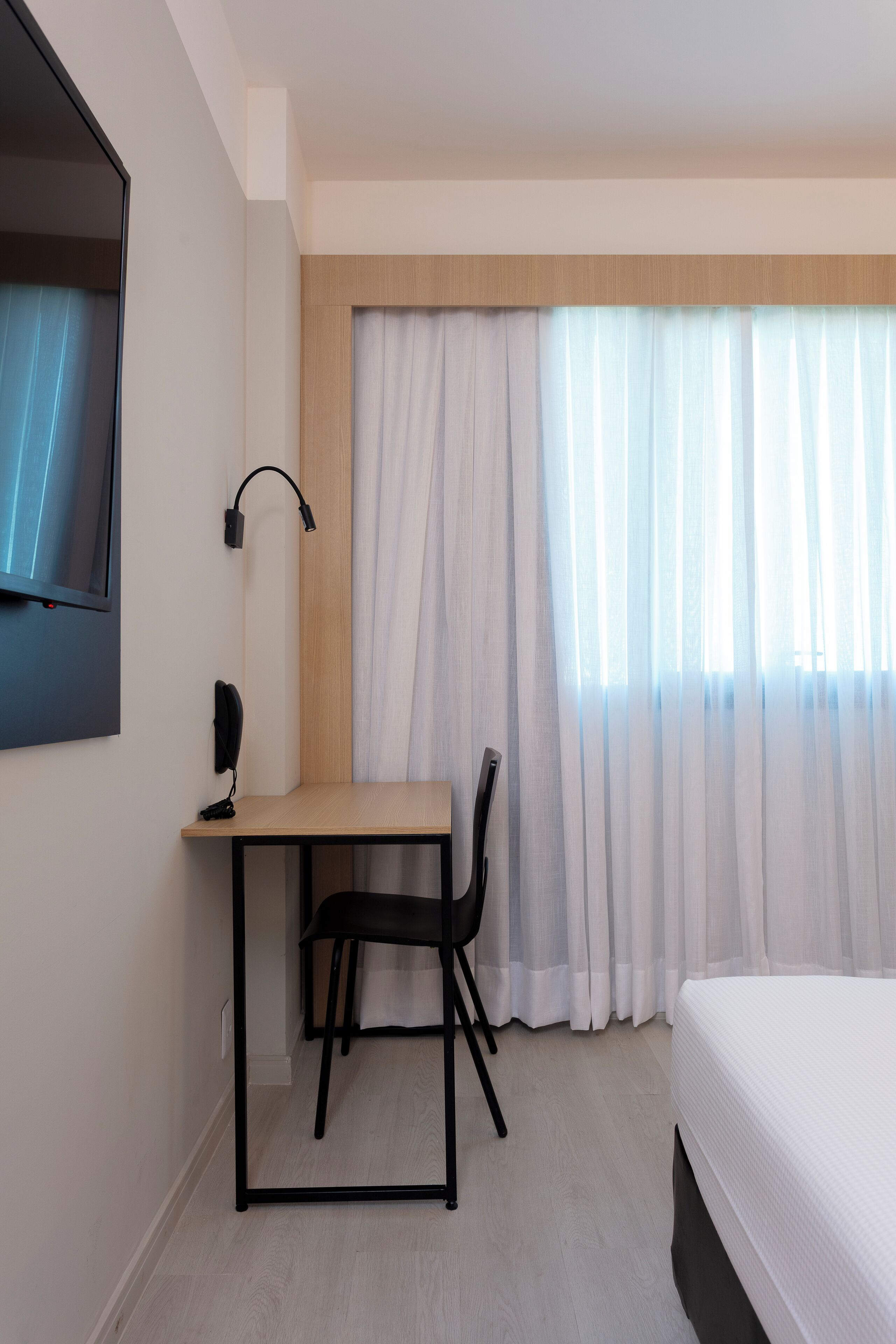 Habitación de hotel con cama, escritorio con silla, televisor montado en la pared y ventana con cortinas traslúcidas.