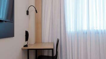 A hotel room with a bed, a desk with a chair, a TV mounted on the wall, and a window with sheer curtains.
