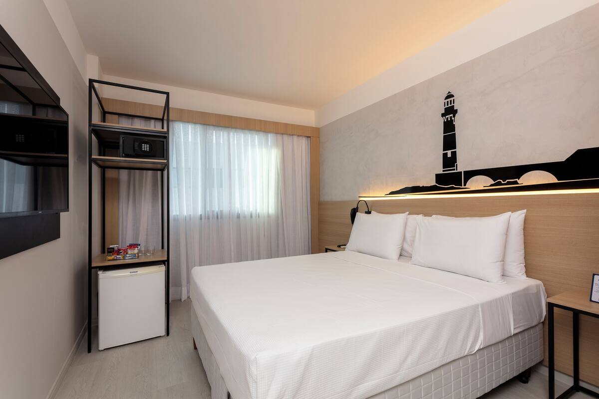 A hotel room with a bed, a desk with a computer, a mini-fridge, and a wall-mounted TV.
