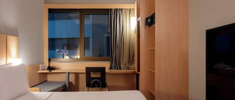 Habitación de hotel con cama, escritorio, silla, televisión y ventana con cortinas.