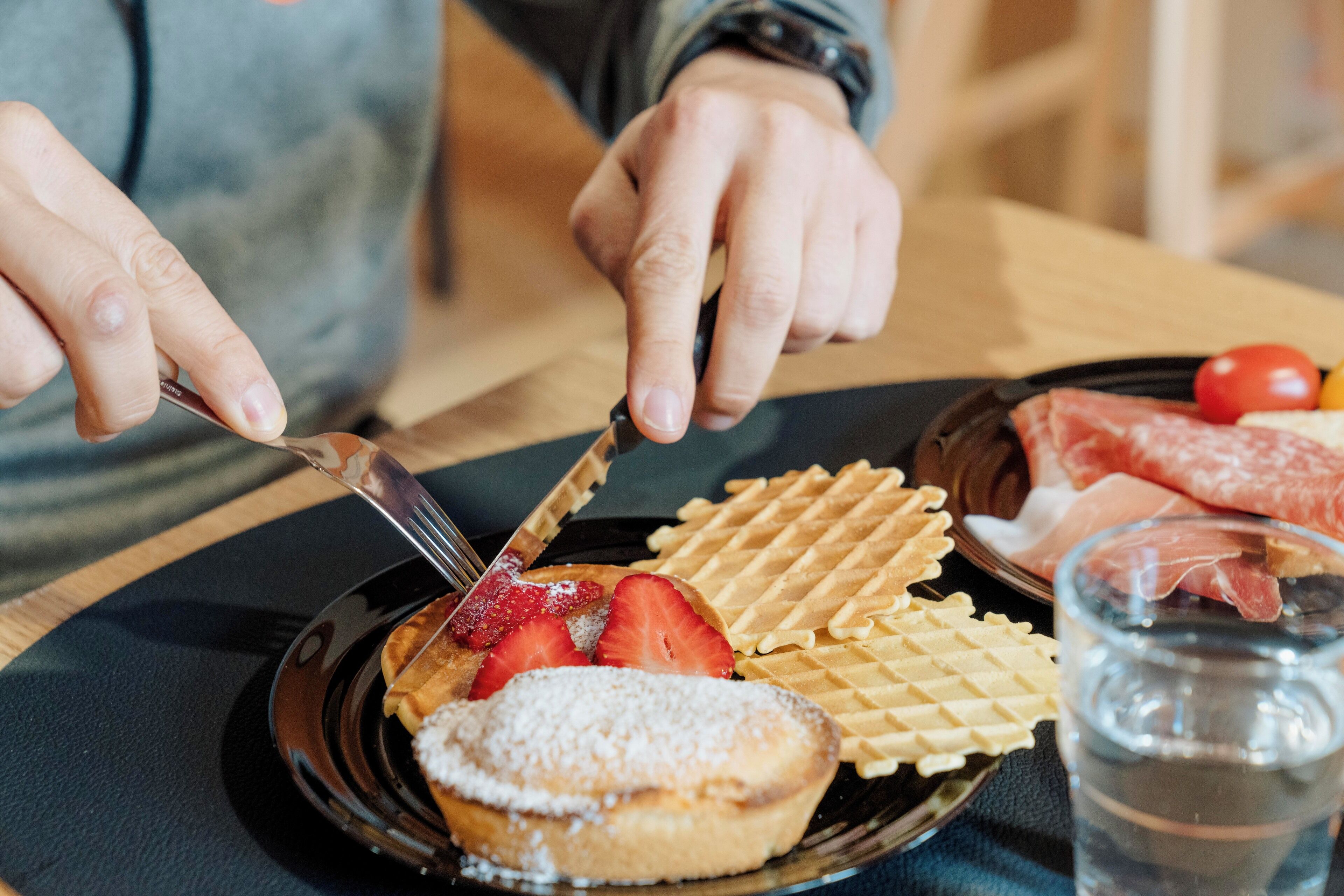 Una persona cortando un gofre con un cuchillo, rodeada de fresas y un pastel en un plato.
