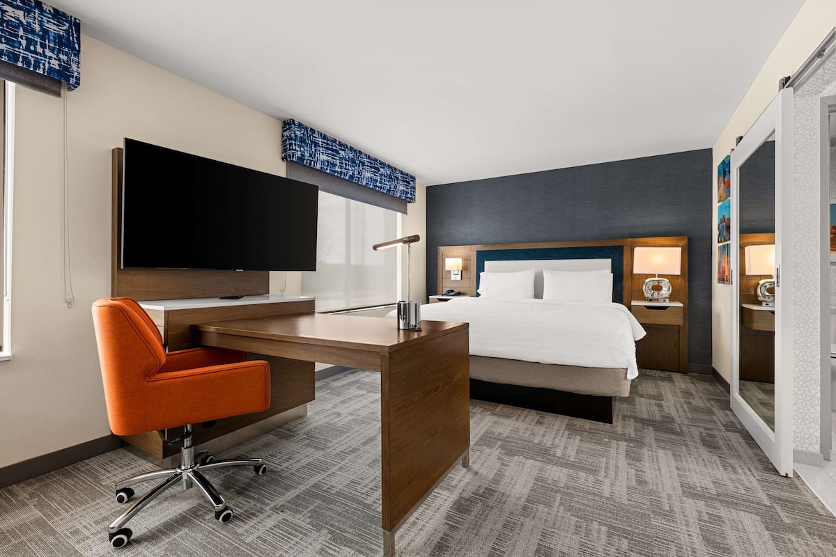 A hotel room with a bed, desk, chair, flat-screen TV, bedside lamps, and a large window.