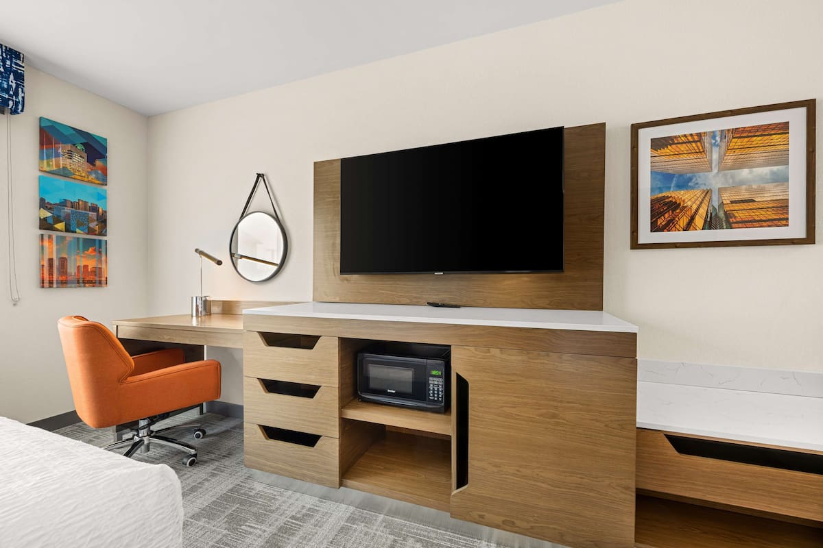 A modern hotel room with a wooden desk, a flat-screen TV, a microwave, and a chair.
