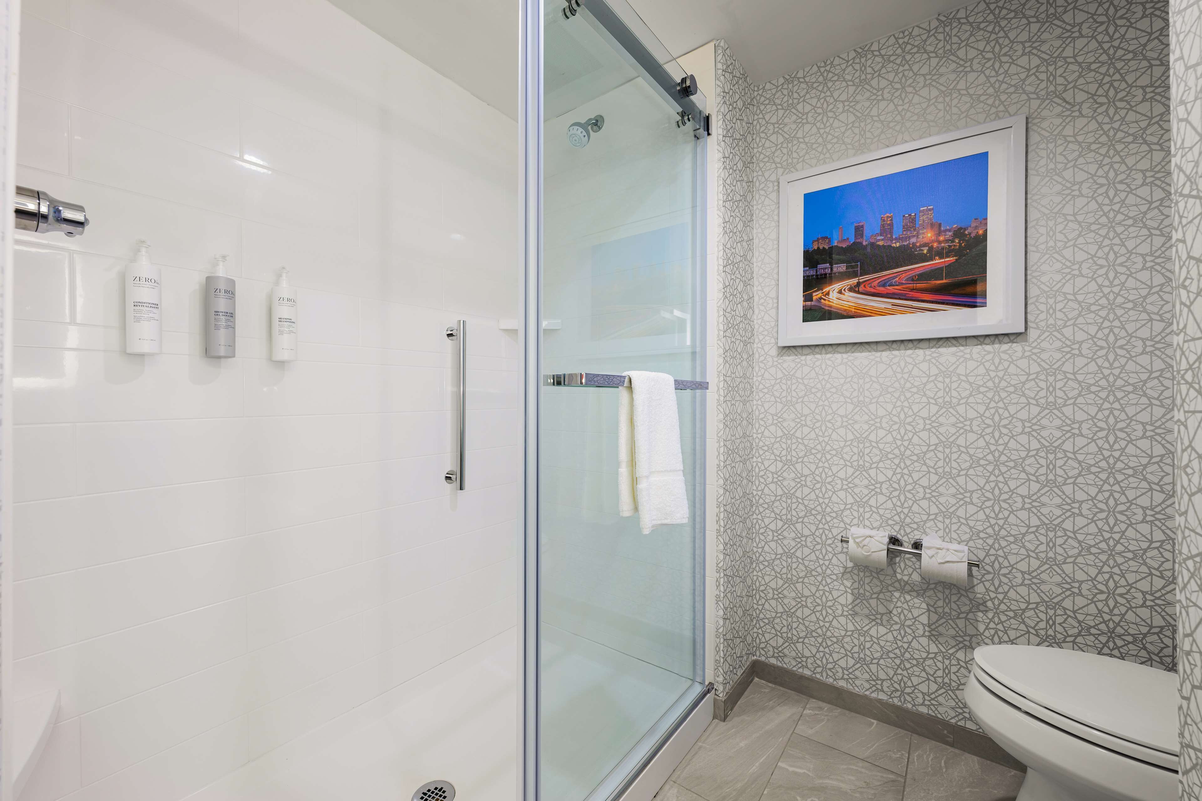 Casa de banho moderna com cabine de duche em vidro, sanita e quadro com uma vista urbana emoldurada na parede.