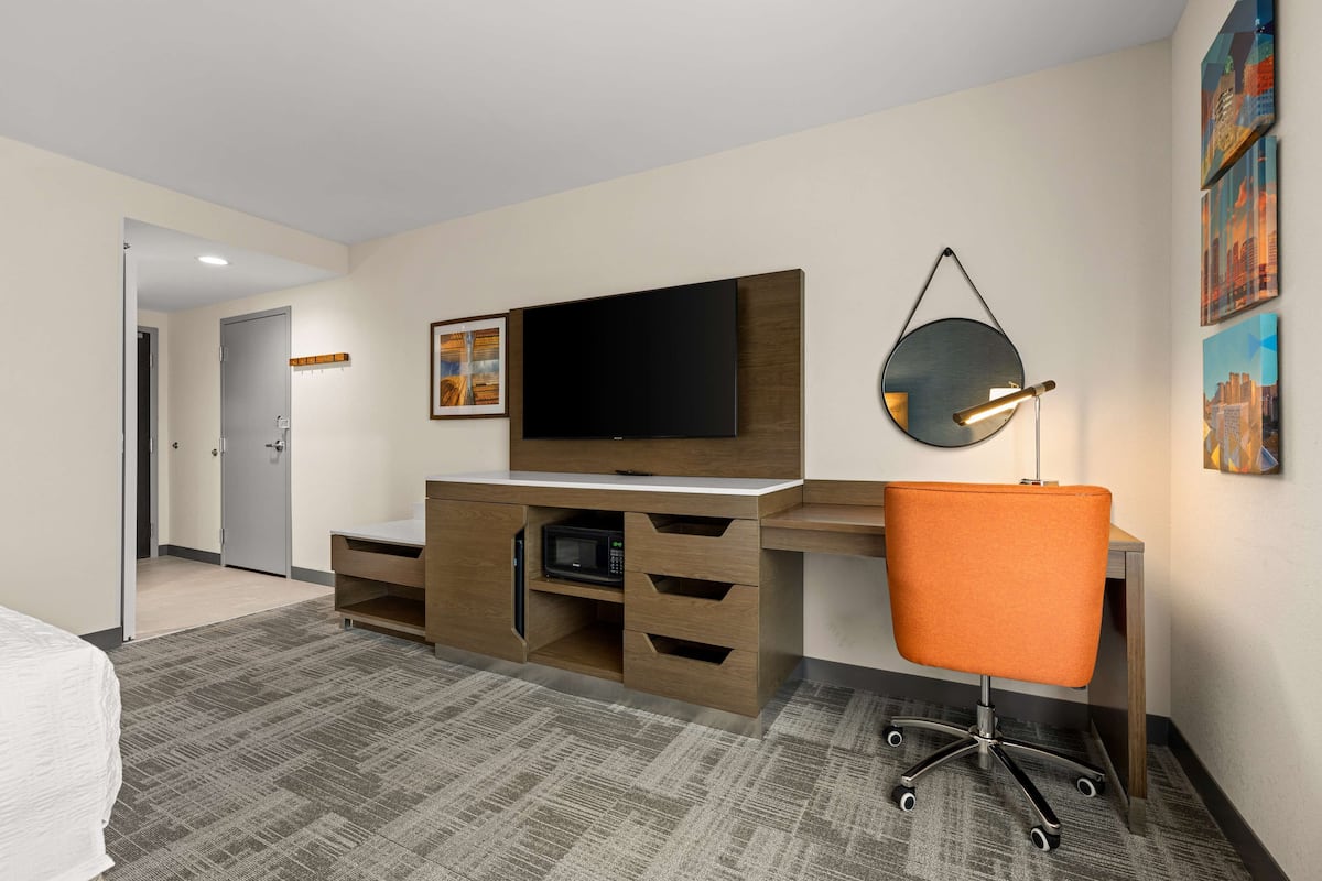 A hotel room with a wooden desk, a flat-screen TV, a microwave, an orange office chair, and a cityscape artwork on the wall.