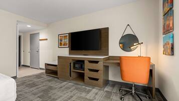 Habitación de hotel con escritorio de madera, televisor de pantalla plana, microondas, silla de oficina color naranja y una obra de arte con un paisaje urbano en la pared.